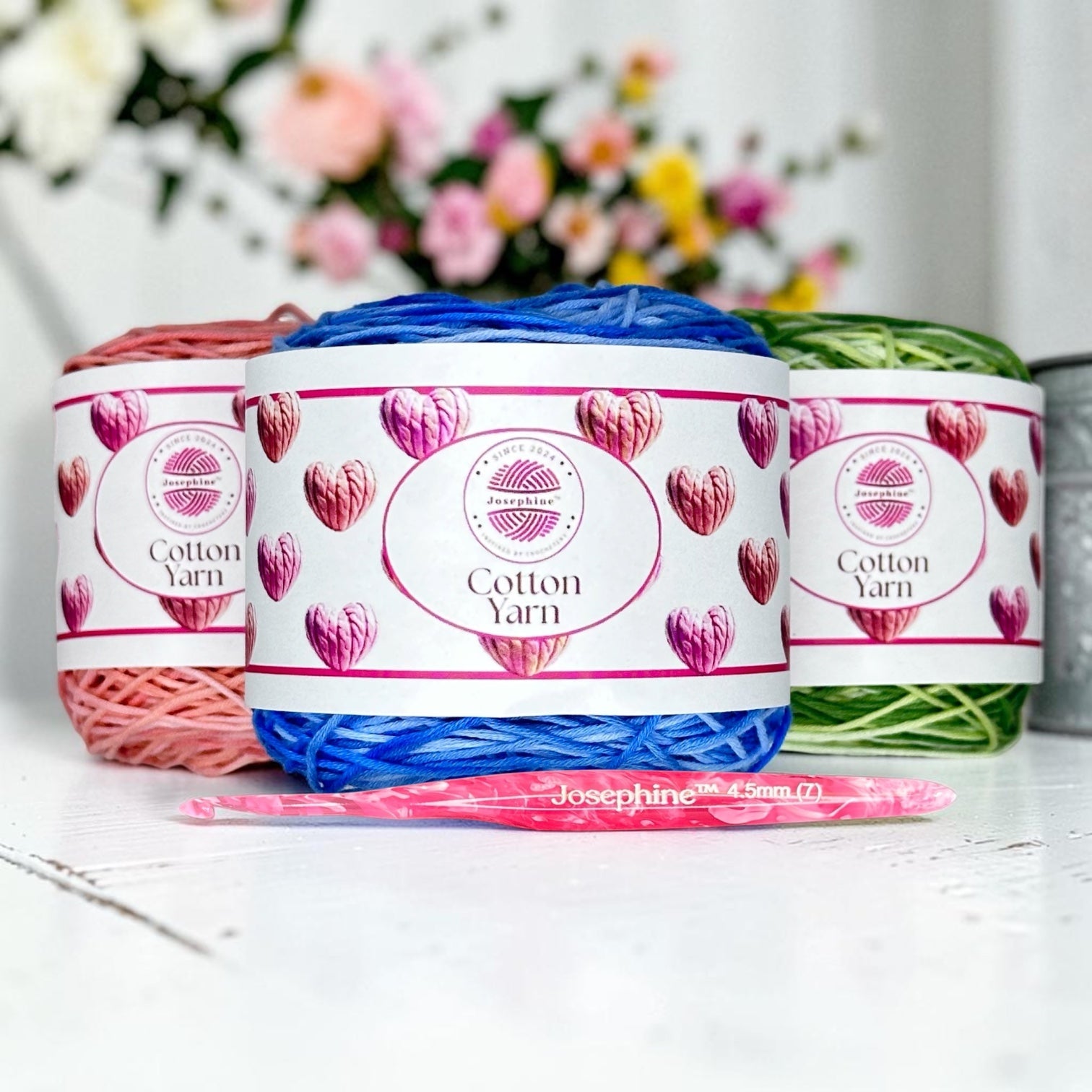 Three skeins of yarn and a Josephine crochet hook with flowers in the background.